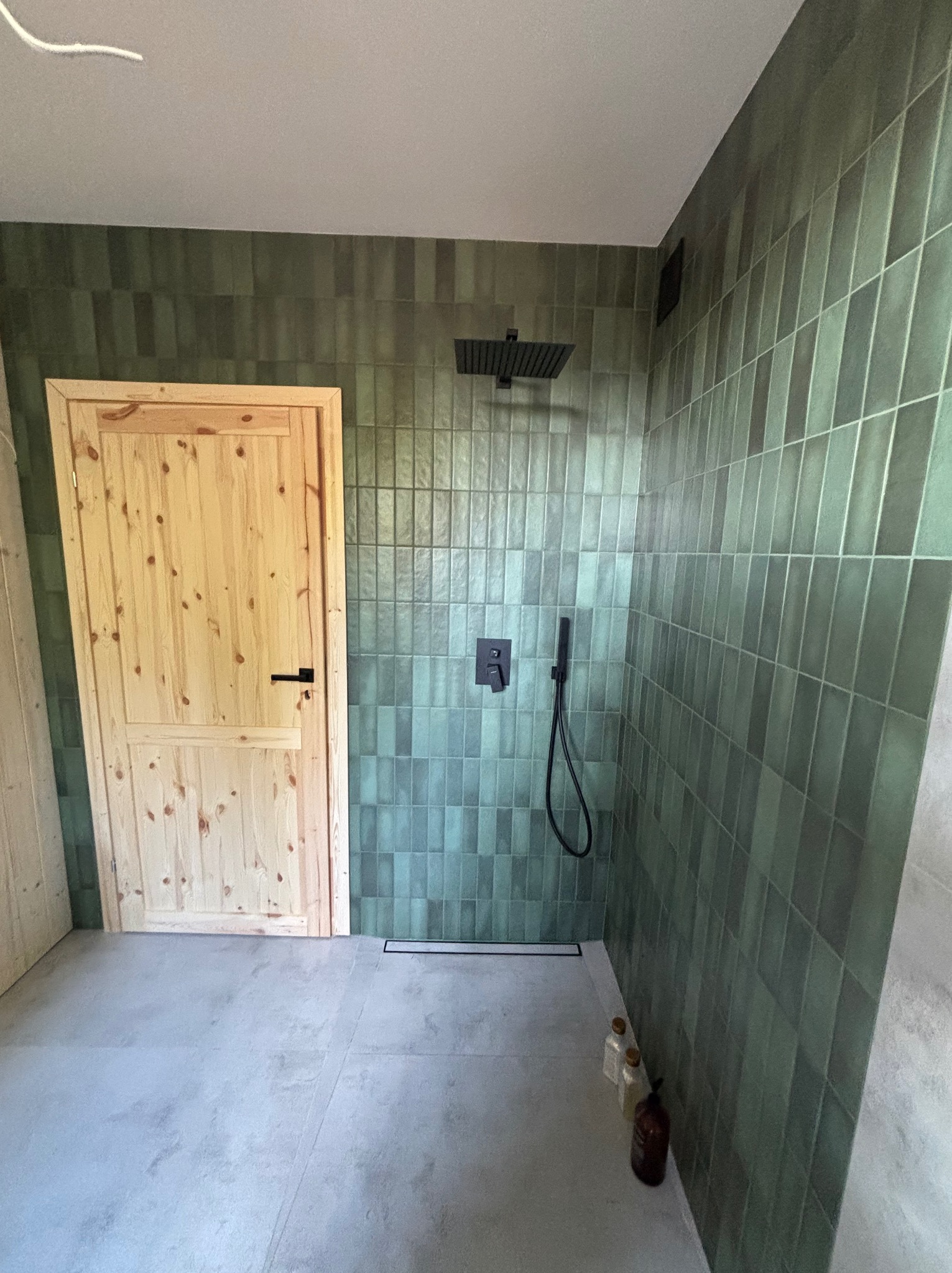 Wykończona łazienka z zielonymi, podłużnymi płytkami, czarną armaturą i drewnianymi drzwiami. Widoczny odpływ liniowy i butelki z kosmetykami.