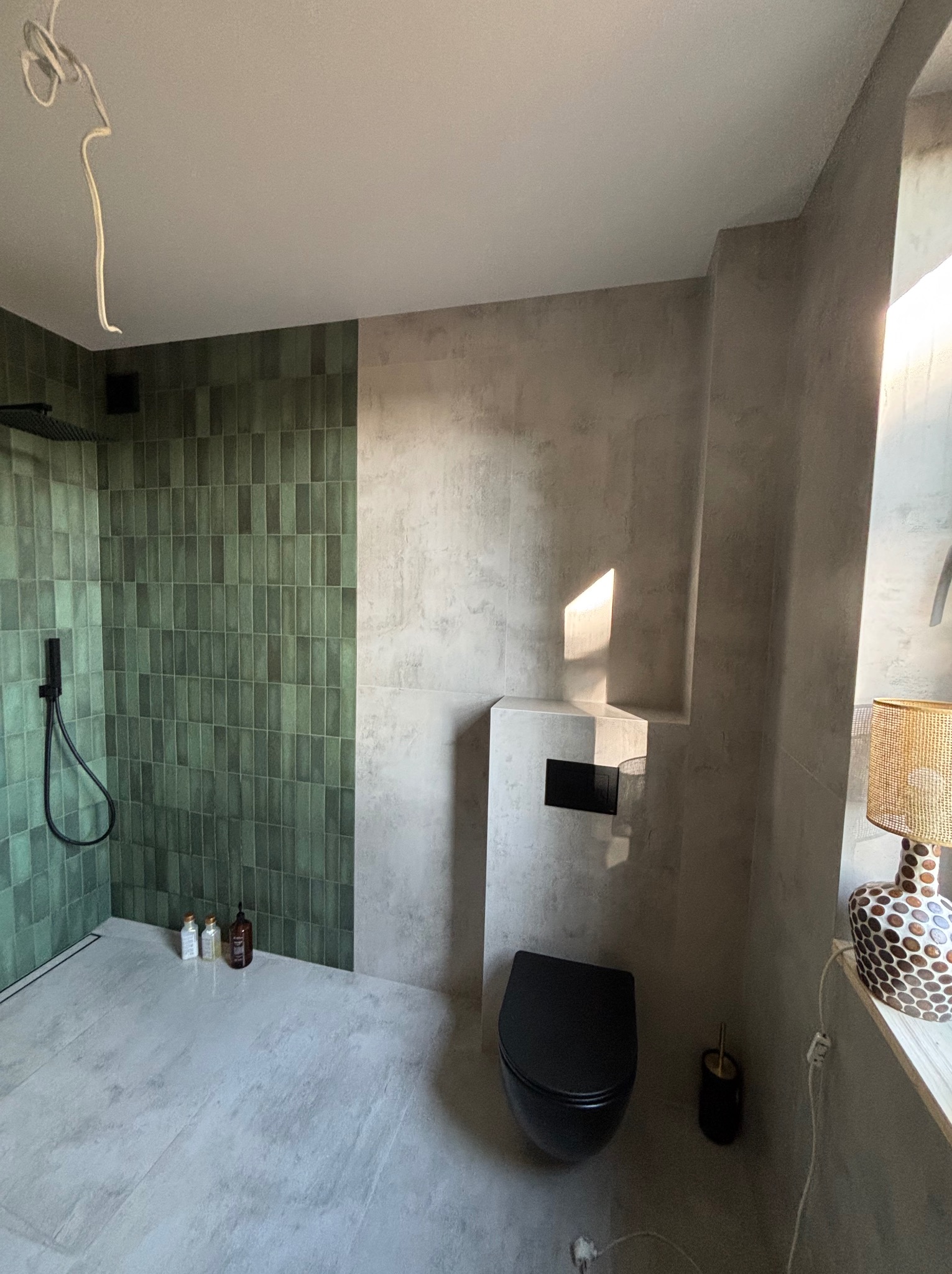 Nowoczesna łazienka z zielonymi płytkami w strefie prysznica i szarą ścianą z podtynkową toaletą. Minimalistyczny design, oświetlenie punktowe i dekoracyjna lampa.