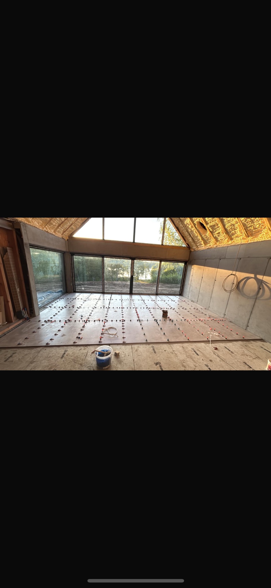 Układanie płytek podłogowych w przestronnym wnętrzu z dużymi oknami. Widoczne poziomice, krzyżyki dystansowe i wiadro z klejem. Przygotowanie do fugowania.