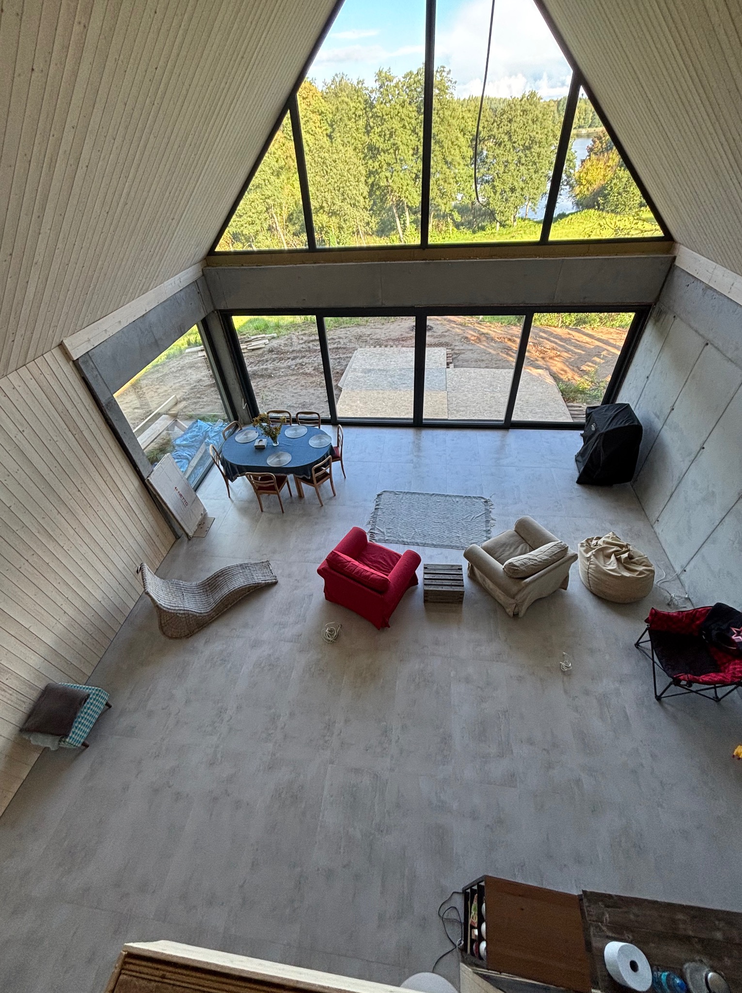 Wysoki kadr wnętrza domu w kształcie litery A z dużymi oknami, betonowymi ścianami i drewnianym wykończeniem sufitu. Widoczny stół, krzesła i elementy dekoracyjne.