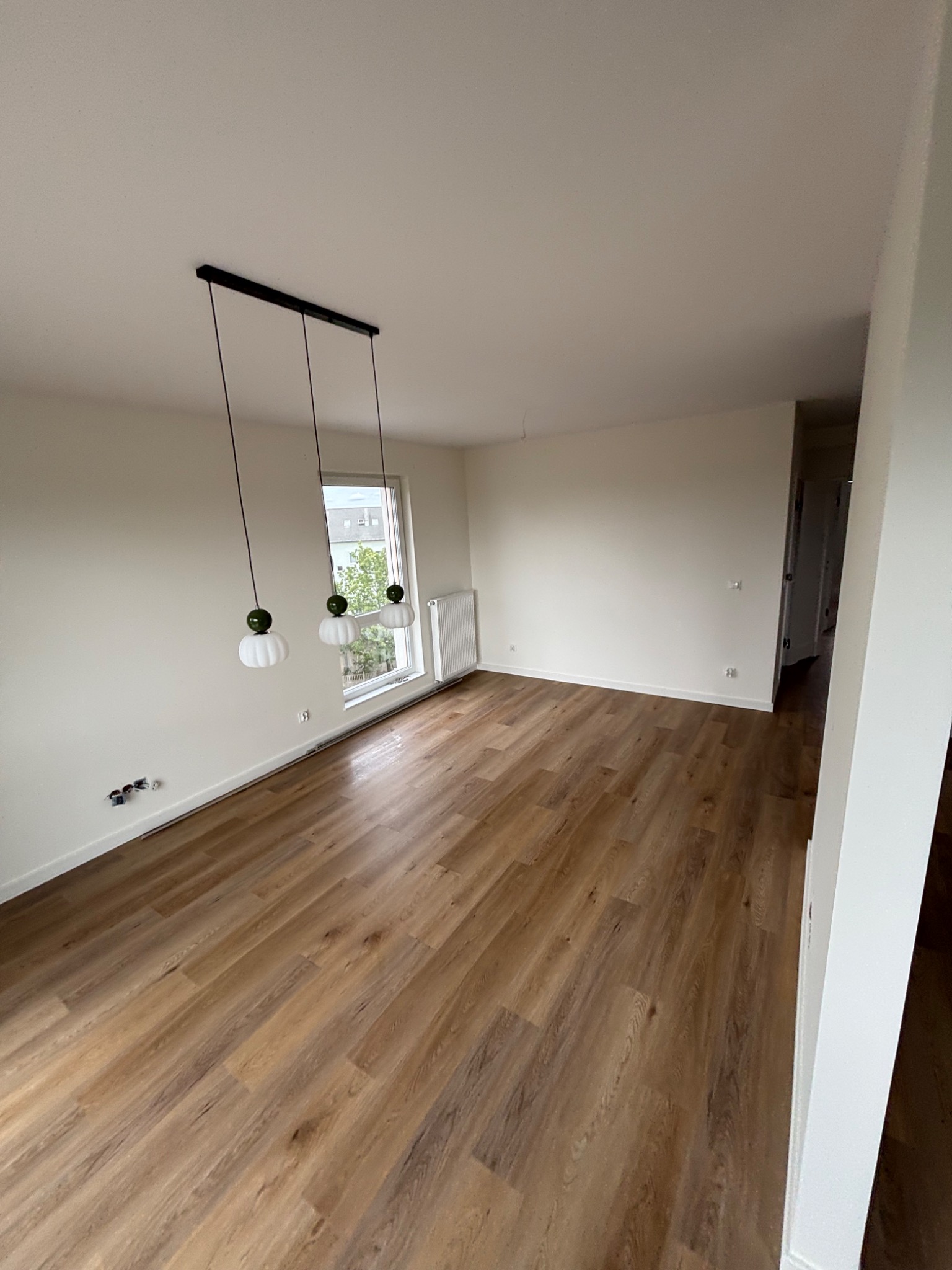 Jasne, przestronne wnętrze po remoncie: drewnopodobna podłoga, świeżo pomalowane ściany, nowoczesna lampa wisząca. Widoczne gniazdka elektryczne i grzejnik.