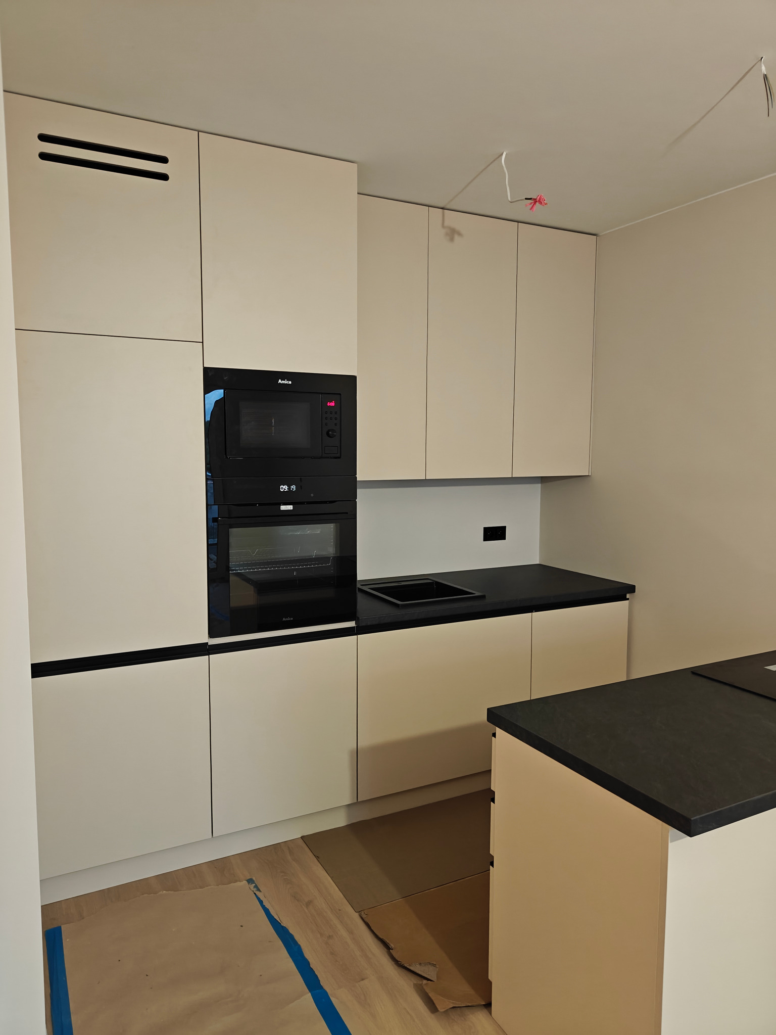 Nowoczesna kuchnia z beżowymi frontami, czarnym blatem i sprzętem Amica. Na podłodze zabezpieczenie z kartonu. Widoczne niedokończone podłączenia elektryczne.