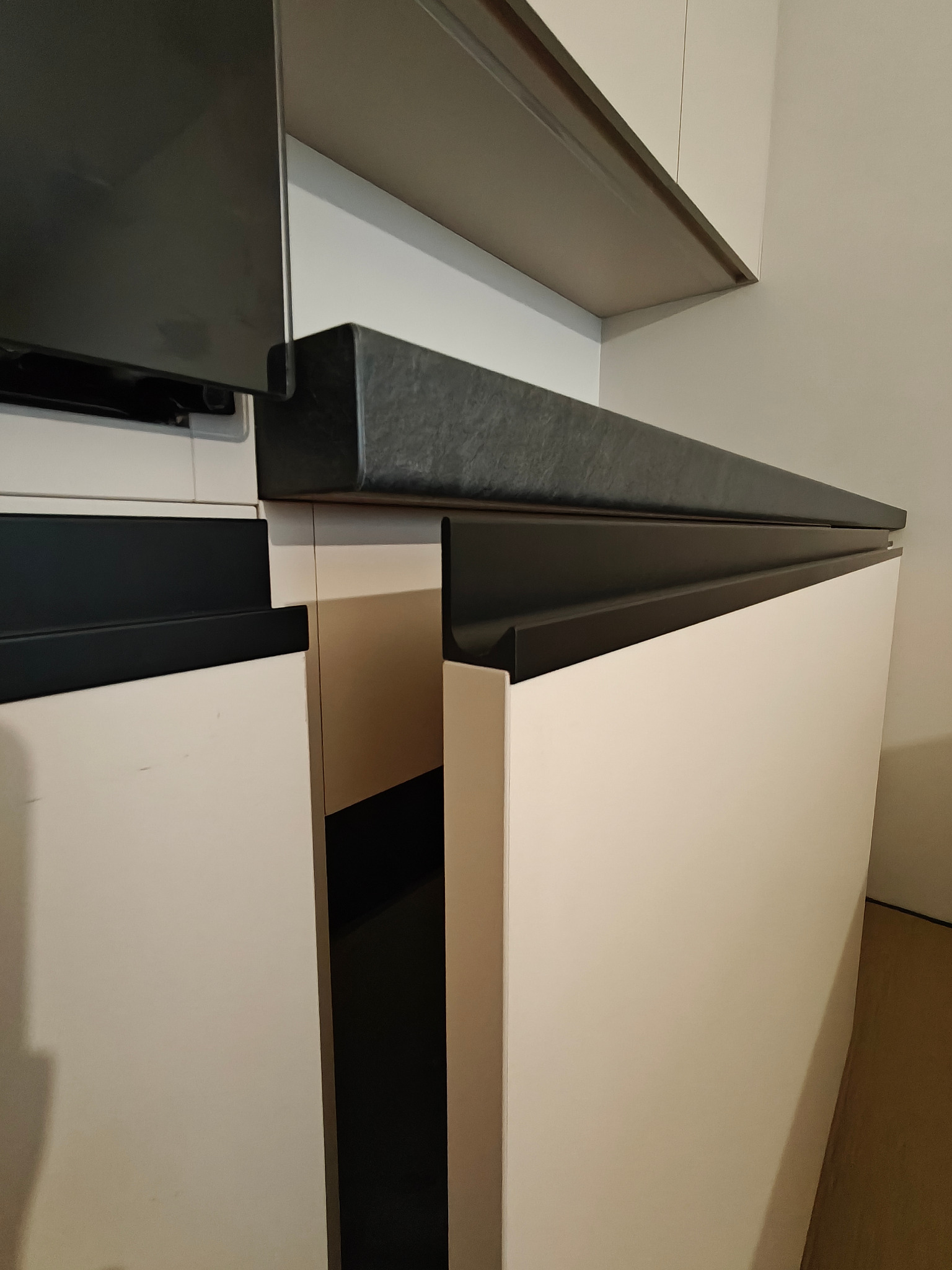 Detale nowoczesnej zabudowy kuchennej: białe fronty szafek z czarnymi, geometrycznymi detalami i blatem, ujęte z bliska pod kątem, z widoczną półką.