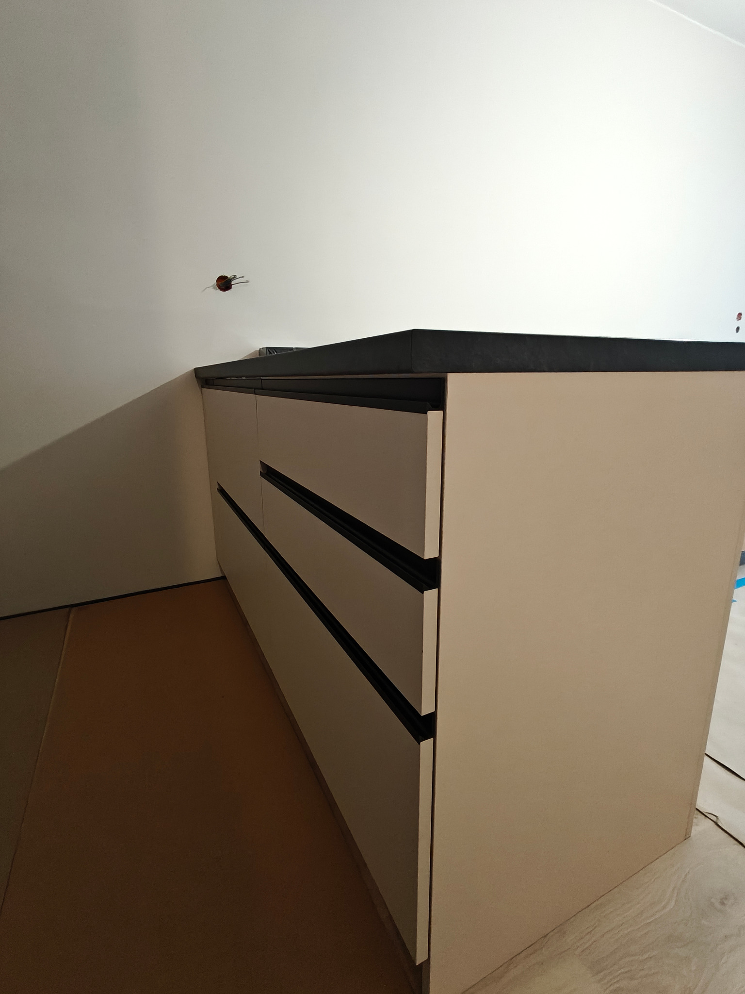 Beżowa szafka z ciemnym blatem i czarnymi uchwytami szuflad, stojąca na zabezpieczonej podłodze, z widocznym gniazdkiem elektrycznym na ścianie.