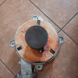 I.S GAS SERWIS SPÓŁKA Z OGRANICZONĄ ODPOWIEDZIALNOŚCIĄ - Zdemontowany palnik gazowy z wyraźnymi śladami użytkowania, osadzony na brązowych płytkach podłogowych. Widoczne elementy mocujące i przewód.