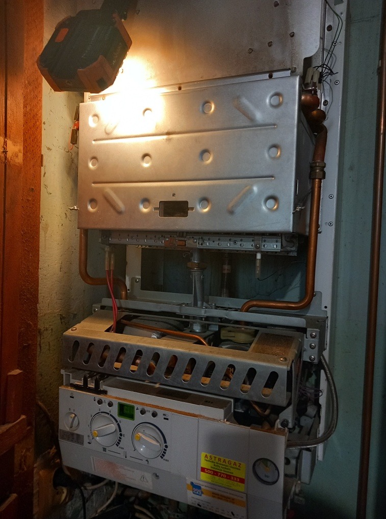 Otwarty piec gazowy marki Astragaz z widocznymi elementami wewnętrznymi i miedzianymi rurami, oświetlony przenośną lampą roboczą.