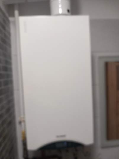 Biały piec gazowy wiszący na ścianie w łazience z szarą, ceglaną ścianą w tle. Widoczny fragment rury. Ujęcie z dołu, lekko rozmyte.