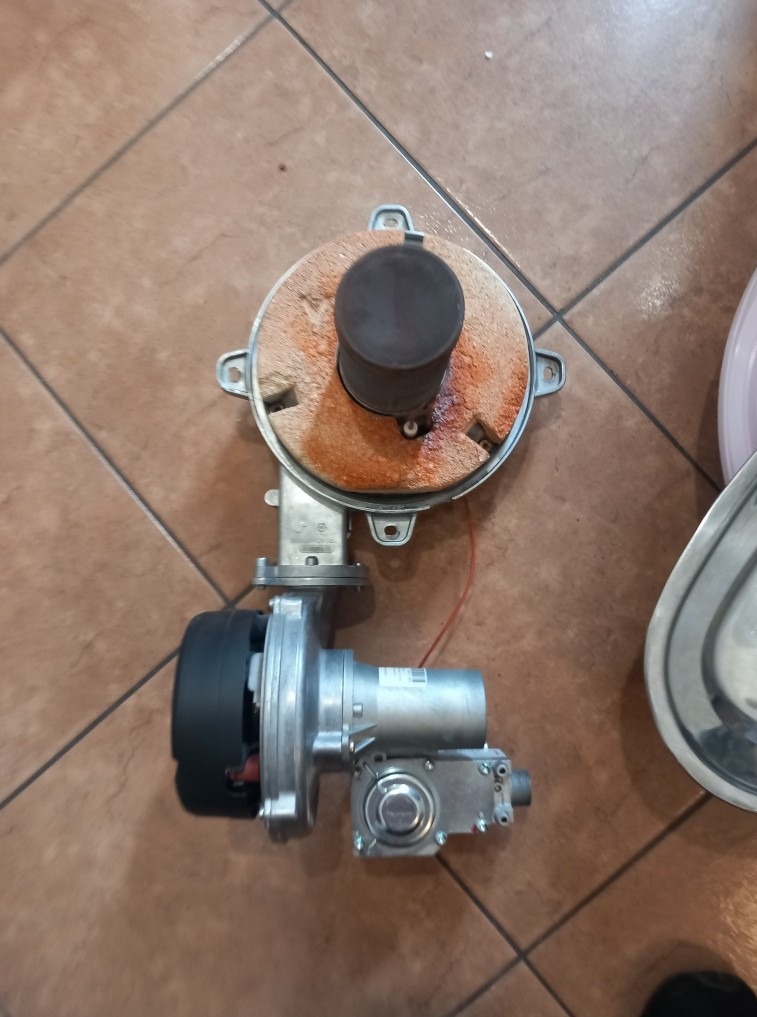 Zdemontowany palnik gazowy z widocznymi śladami użytkowania leży na brązowych płytkach. Elementy są metalowe, z widoczną rdzą i nagarem. Po prawej widoczny fragment naczynia.