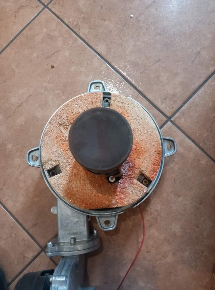 Zdemontowany palnik gazowy z wyraźnymi śladami użytkowania, osadzony na brązowych płytkach podłogowych. Widoczne elementy mocujące i przewód.