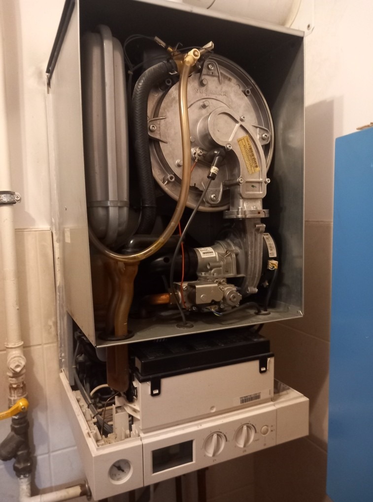 Otwarty piec gazowy Viessmann, widoczne wewnętrzne elementy, rury i przewody. Biała obudowa, płytki w tle. Układ pionowy.