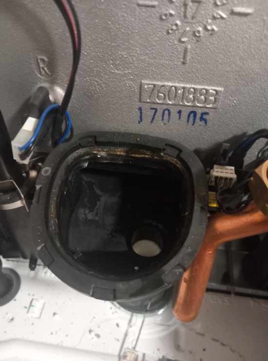 Otwarty, czarny element pieca gazowego z widocznymi numerami seryjnymi i fragmentami instalacji miedzianej. Detal naprawy lub przeglądu kotła gazowego.