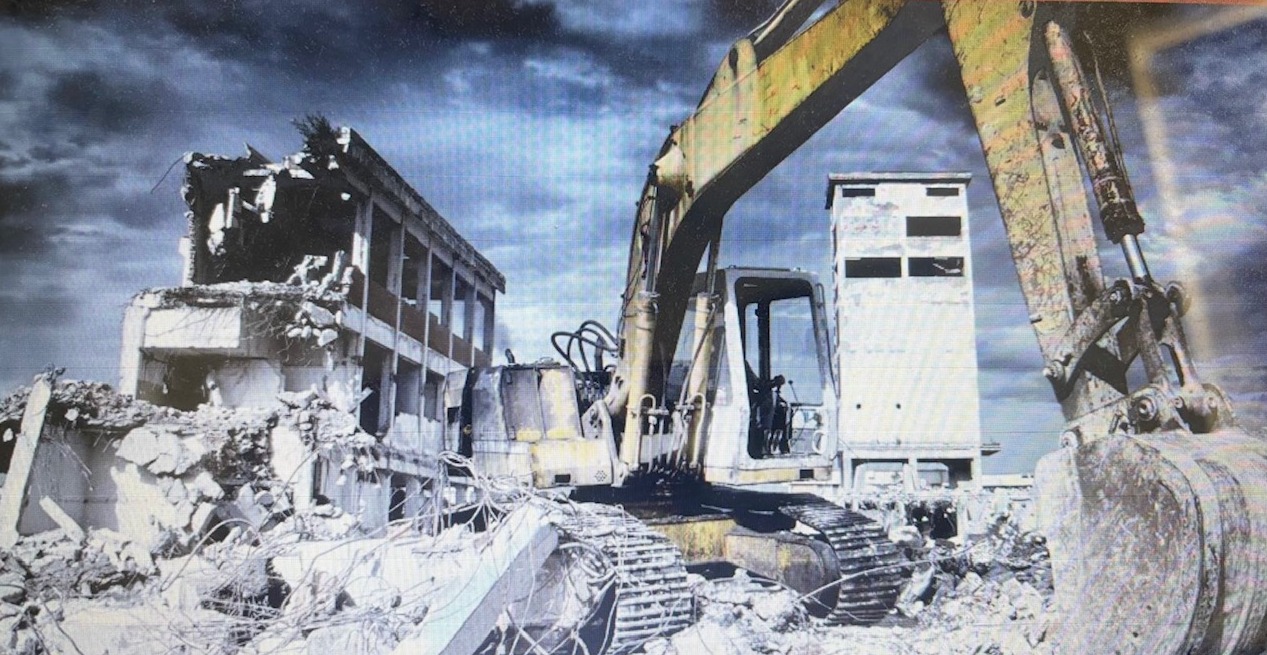 Żółta koparka na gruzach budynku, rozbiórka w toku. Fragment konstrukcji i ponure, zachmurzone niebo w tle. Prace rozbiórkowe.