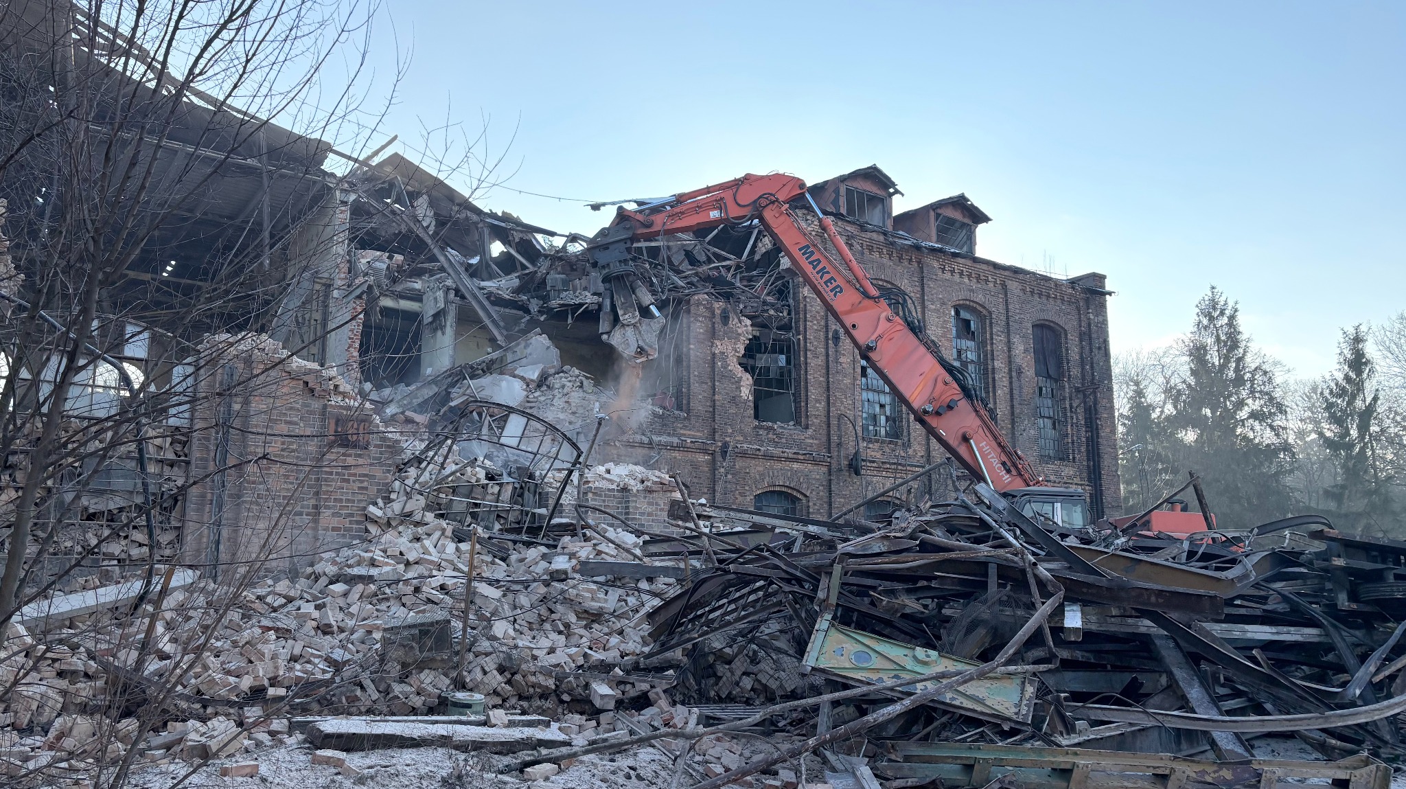 Częściowo zburzony ceglany budynek z pomarańczowym ramieniem koparki Maker, gruz i elementy konstrukcyjne na pierwszym planie, drzewa w tle.