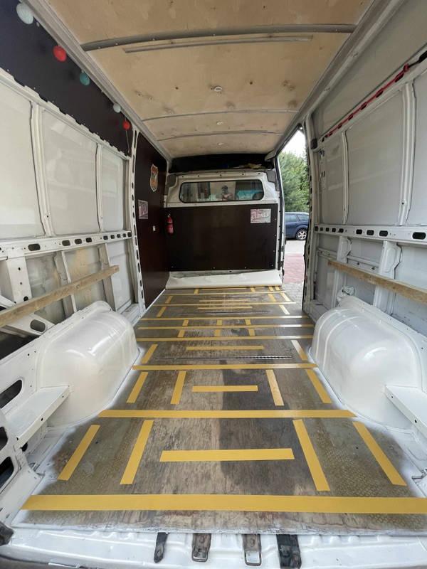 Wnętrze dostawczego busa z widoczną podłogą wyłożoną płytami drewnianymi i wzmocnioną żółtymi taśmami. Widoczne boki i sufit przestrzeni ładunkowej.