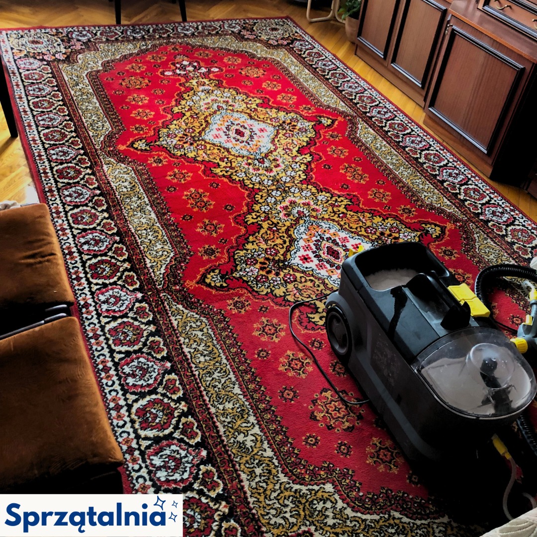 Czerwony, wzorzysty dywan czyszczony odkurzaczem piorącym. Widoczne drewniane nogi krzesła i parkiet. Perspektywa z góry ukazuje detale dywanu i sprzętu.