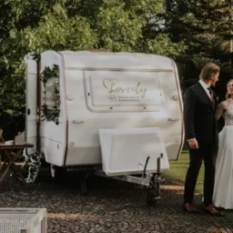 Biała fotobudka w formie przyczepy z napisem 'Loventy' na tle zieleni. Para młoda stoi obok, patrząc na siebie. Dekoracje kwiatowe i drewniane rekwizyty.