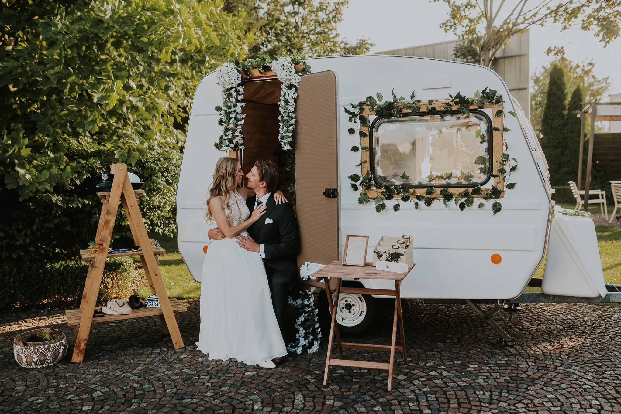 Para młoda pozuje przed białą fotobudką-przyczepą, ozdobioną kwiatami i zielenią. Dekoracje weselne, drewniane rekwizyty, romantyczna atmosfera.