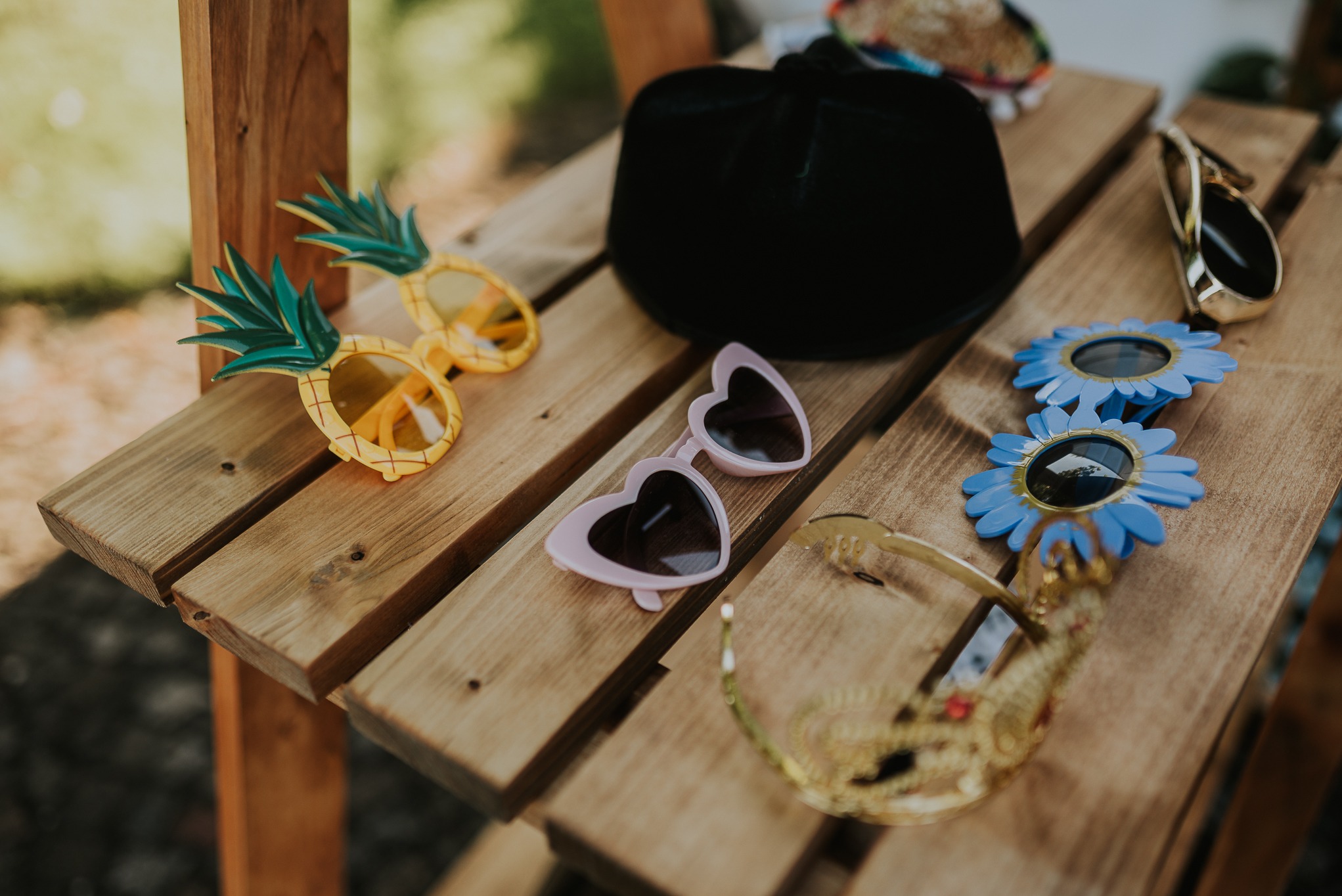 Różne rekwizyty do fotobudki: okulary w kształcie ananasa, serca, kwiatów, złota korona i czarny kapelusz, ułożone na drewnianym stoliku.