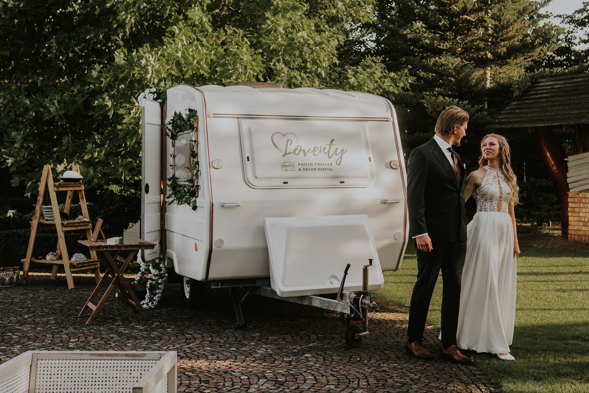 Biała fotobudka w formie przyczepy z napisem 'Loventy' na tle zieleni. Para młoda stoi obok, patrząc na siebie. Dekoracje kwiatowe i drewniane rekwizyty.