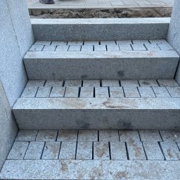 Akvit Siarhei bruk - Schody z jasnoszarej kostki granitowej w trakcie układania, widoczne szczeliny między kostkami, fragmenty obuwia robotników w tle.