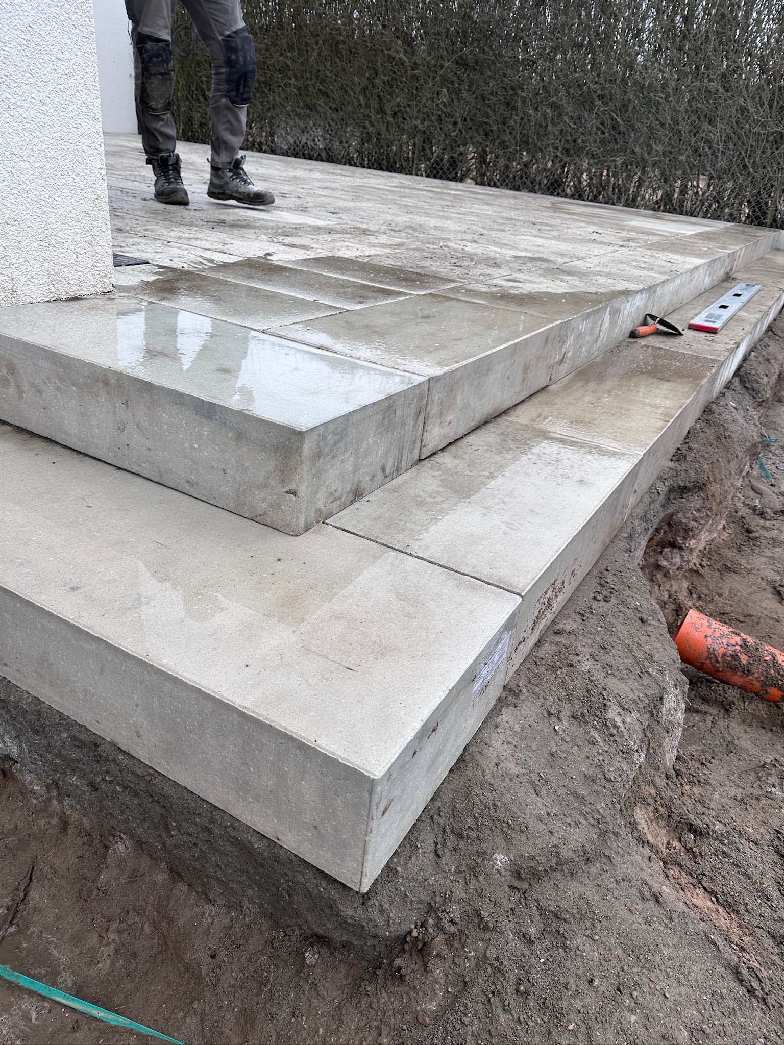 Ułożone stopnie z jasnoszarej kostki. Widoczne narzędzia pomiarowe i fragment instalacji. Częściowo widoczna osoba w stroju roboczym.