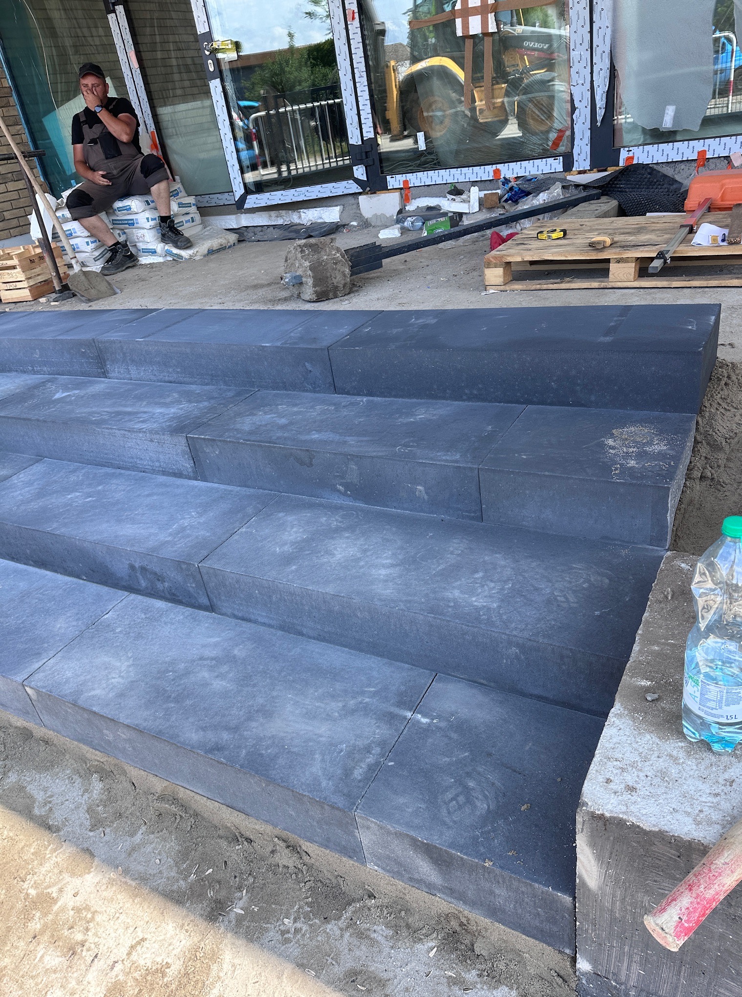 Nowe, ciemnoszare schody z kostki brukowej prowadzą do budynku. W tle pracownik w kombinezonie siedzi na workach cementu. Widoczny sprzęt budowlany i narzędzia.