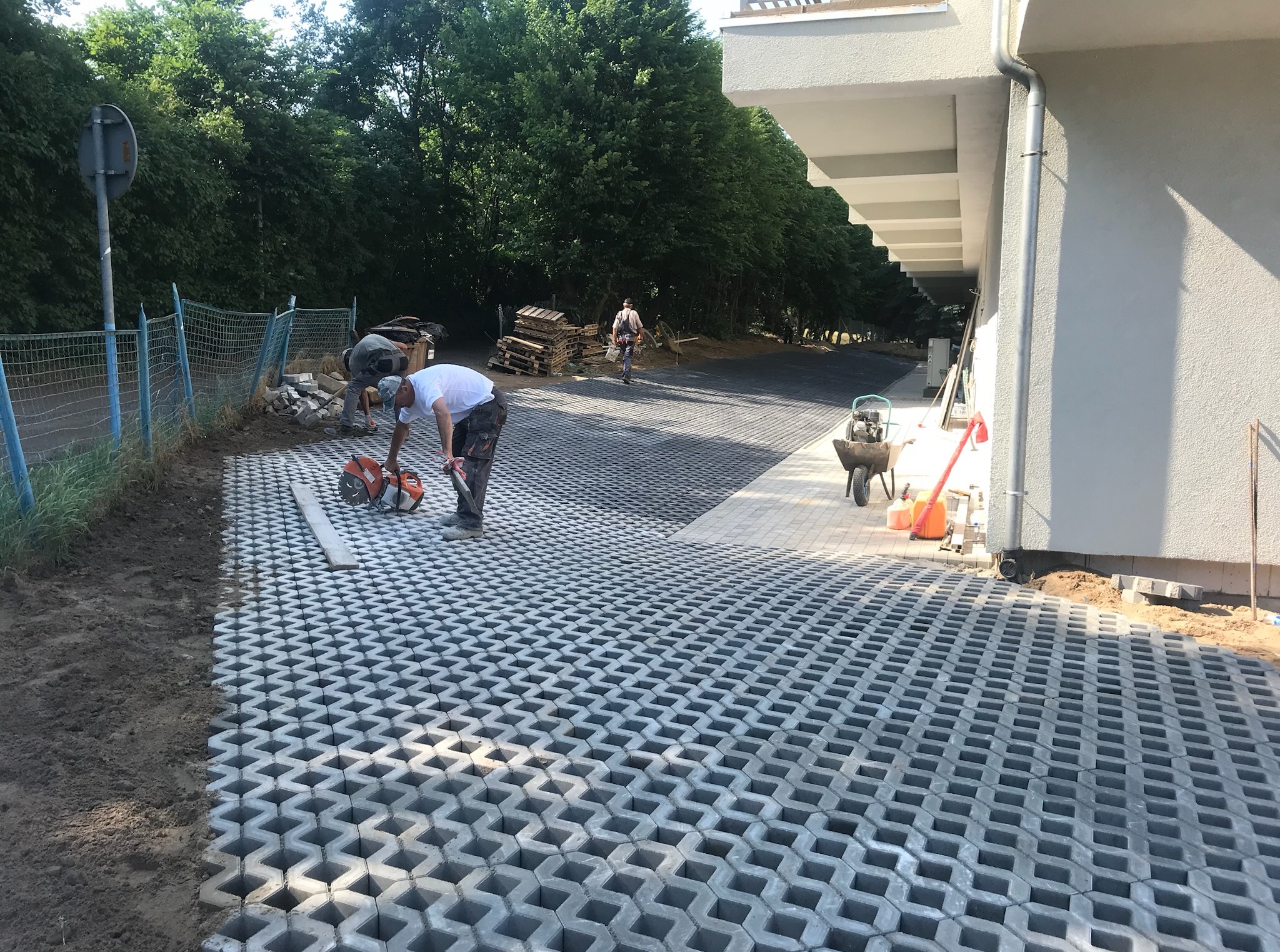 Montaż ażurowej kostki brukowej na posesji. Pracownik docina element. Widoczny budynek, drzewa i narzędzia. Częściowo ułożona nawierzchnia.