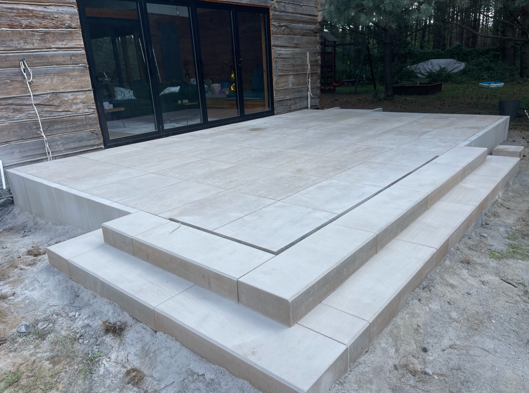 Jasne, kwadratowe płyty tarasowe ułożone schodkowo przed drewnianym domem z dużymi, przesuwnymi drzwiami, otoczone zielenią.