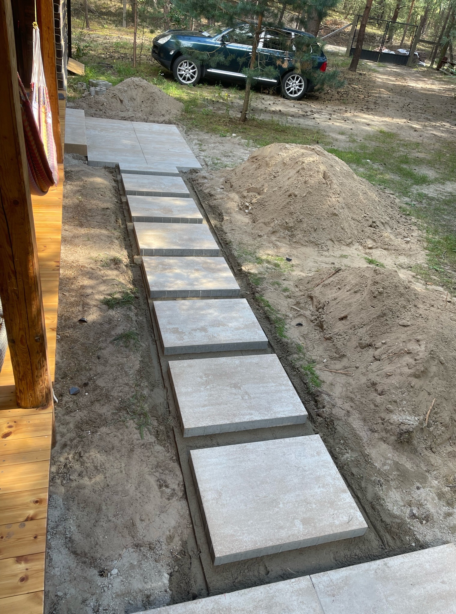 Ścieżka z jasnoszarych płyt betonowych w ogrodzie, prowadząca od drewnianego tarasu w kierunku zaparkowanego, zielonego SUV-a. Widoczne pryzmy piasku.