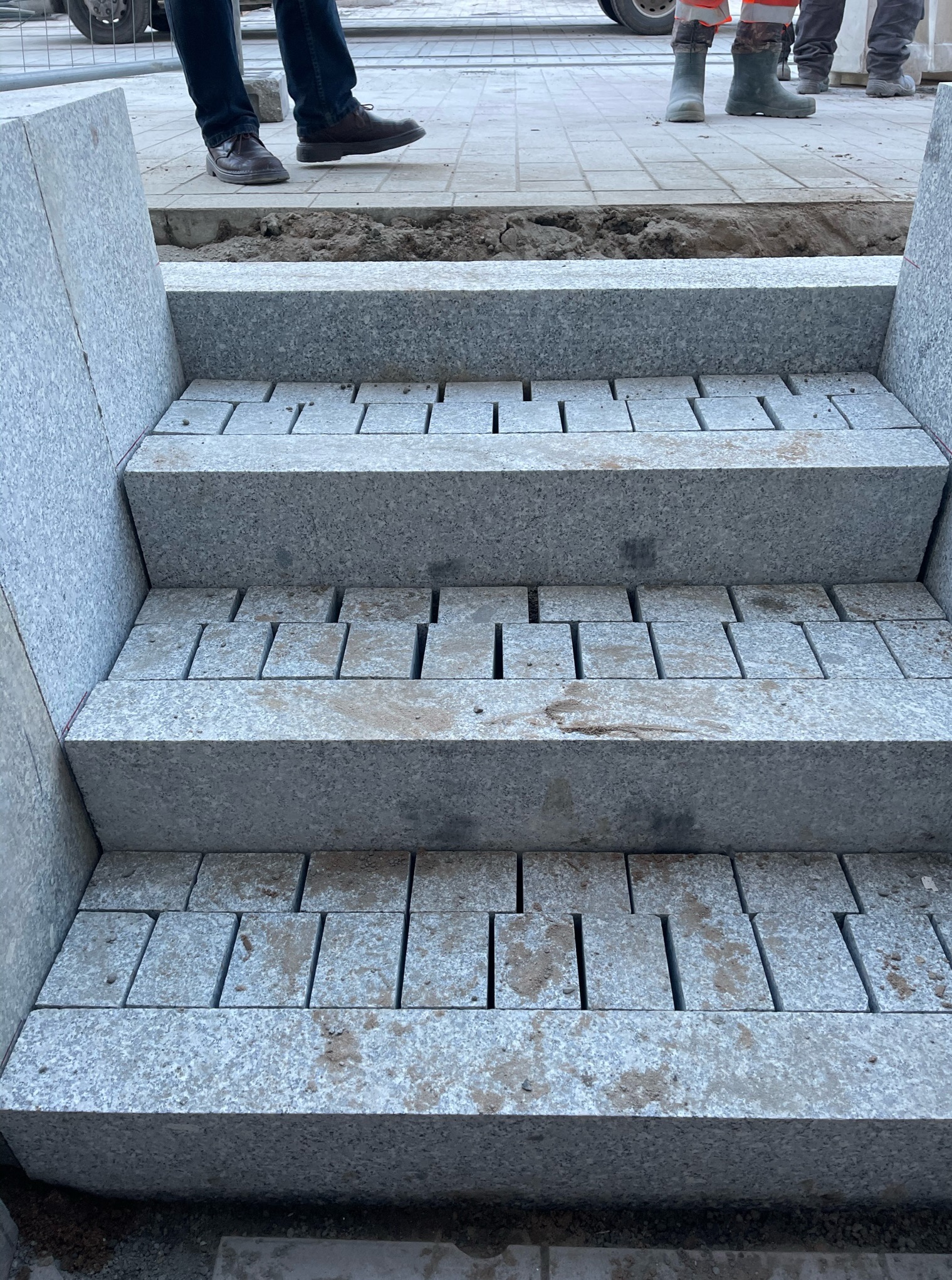 Schody z jasnoszarej kostki granitowej w trakcie układania, widoczne szczeliny między kostkami, fragmenty obuwia robotników w tle.
