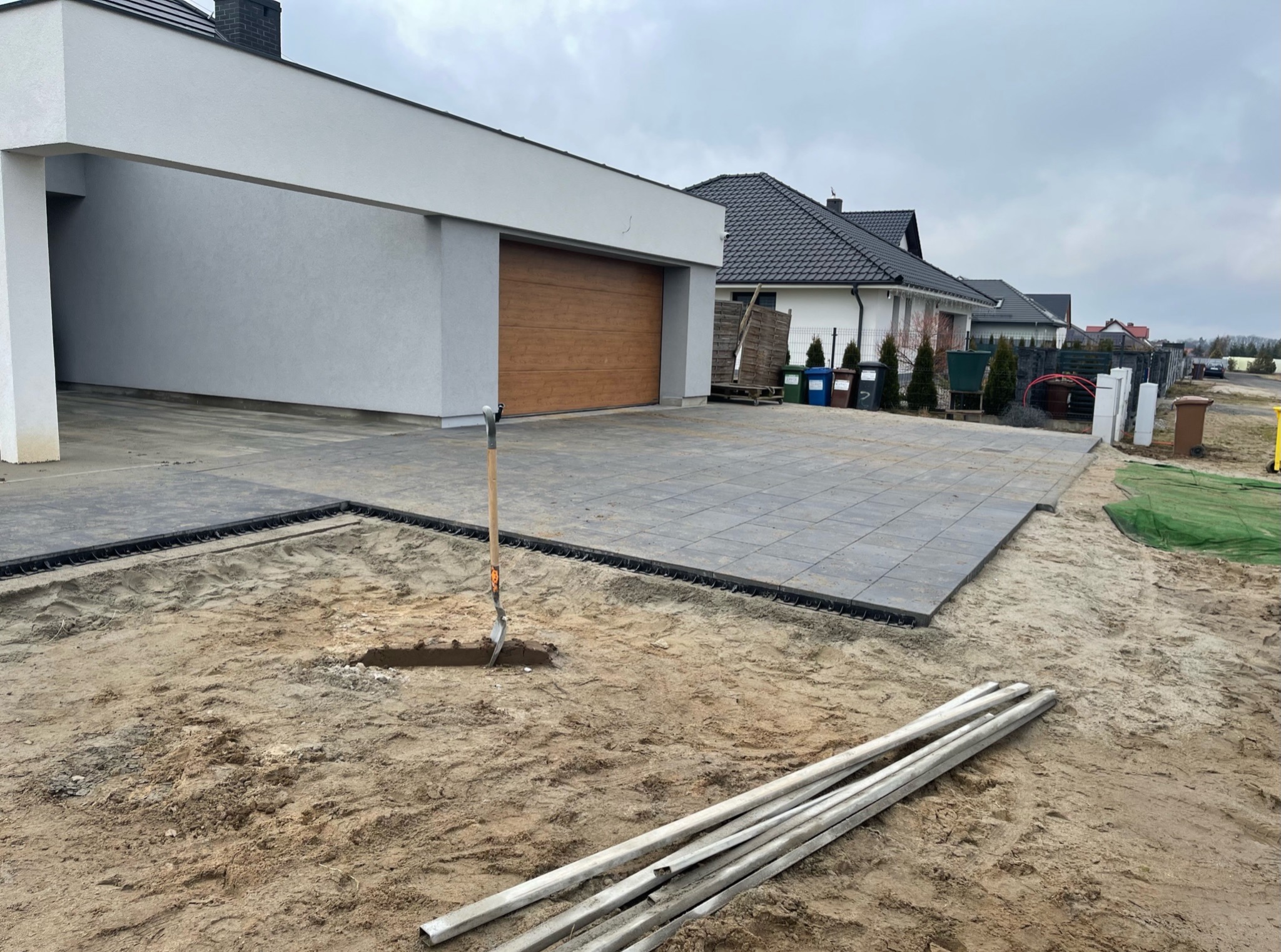 Fragment posesji w trakcie prac brukarskich: ułożona część kostki, wykop pod dalszą część, rury i łopata. Tło: dom z garażem i sąsiednie zabudowania.