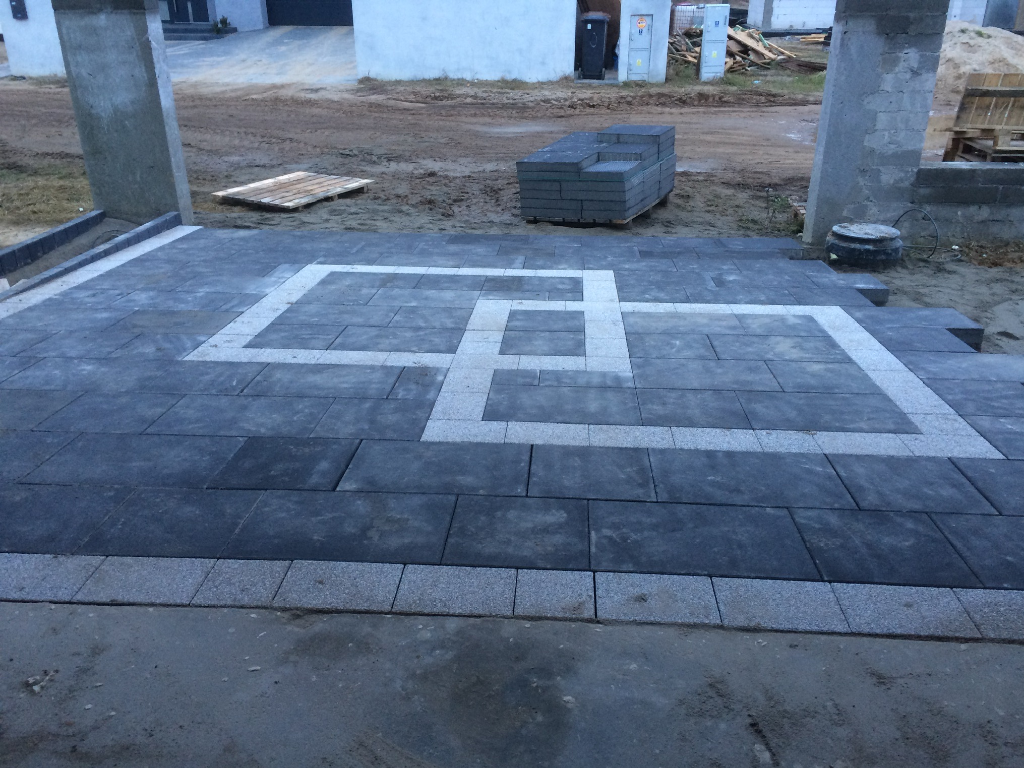 Ułożona kostka brukowa w geometryczny wzór z jasnymi obramowaniami. Widoczne palety z kostką i elementy infrastruktury w tle.