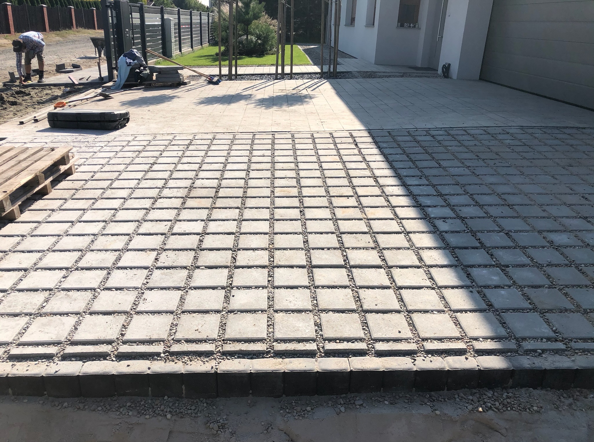 Częściowo ułożona, przepuszczalna nawierzchnia z betonowych płyt i żwiru na podjeździe, z pracującym brukarzem w tle i widocznym fragmentem posesji.