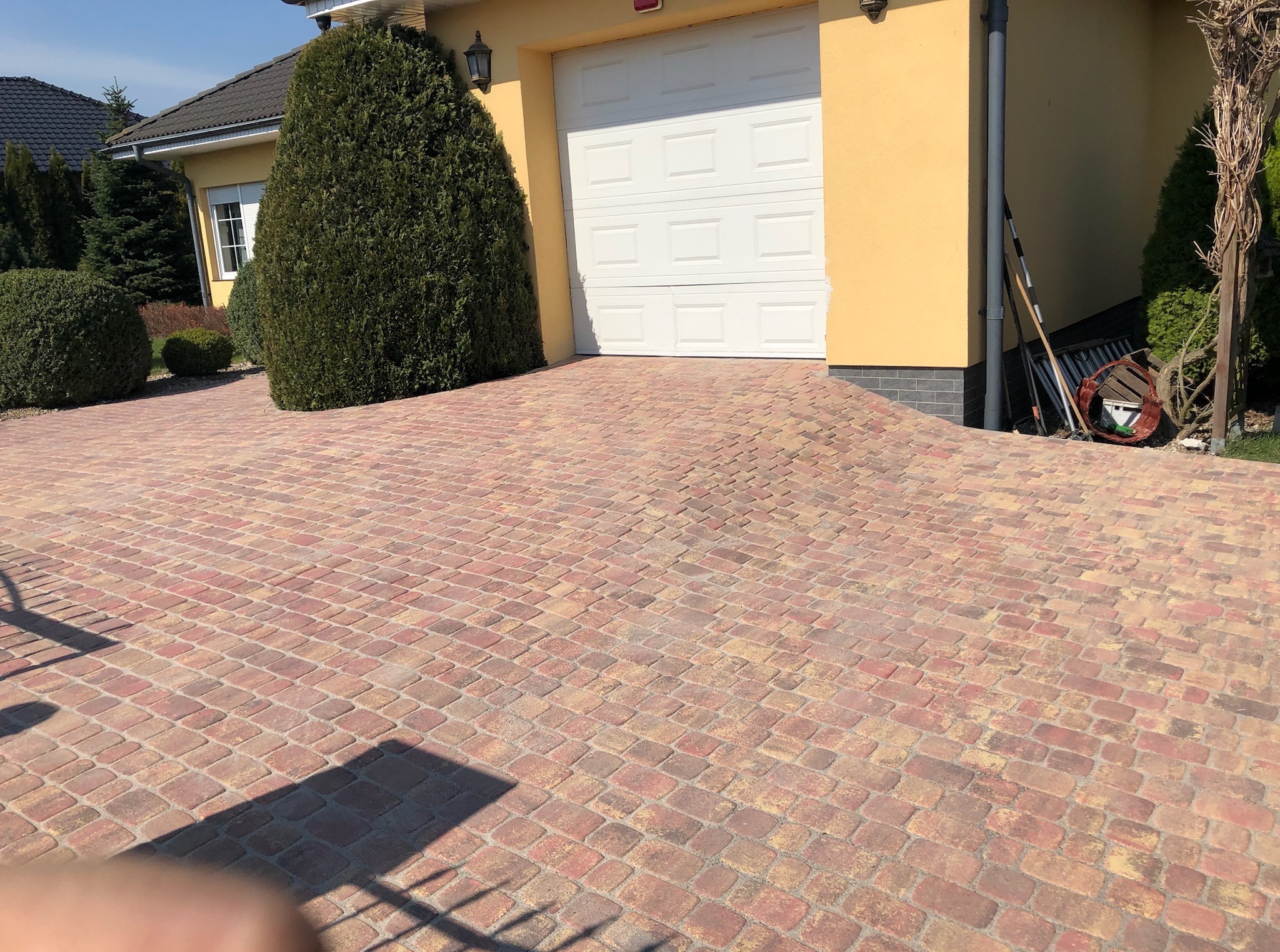 Podjazd z kostki brukowej w odcieniach czerwieni i beżu, prowadzący do garażu w żółtym domu z zadbanym ogrodem. Widoczne narzędzia ogrodnicze.