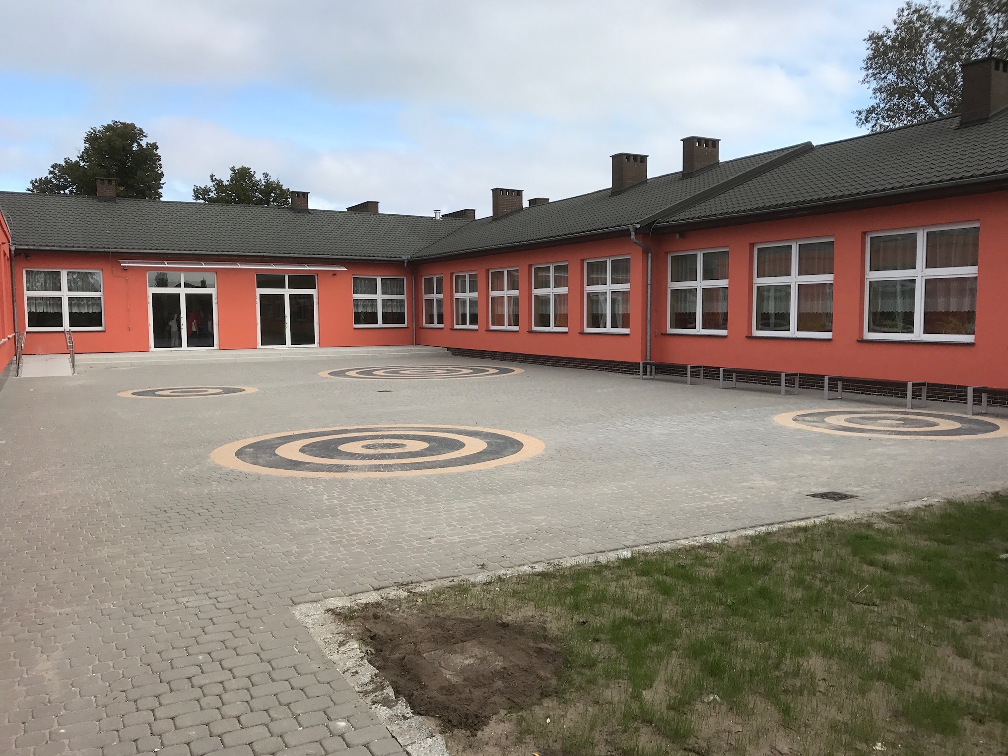 Dziedziniec szkolny z szarej kostki brukowej w okręgi. Budynek w tle pomalowany na pomarańczowo z białymi oknami. Widok z poziomu gruntu.