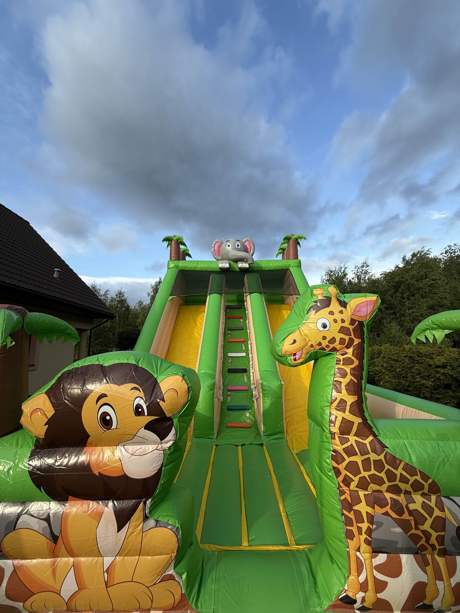 Dmuchana zjeżdżalnia w motywie safari z lwem, żyrafą i słoniem na tle pochmurnego nieba. Atrakcja na imprezę plenerową.