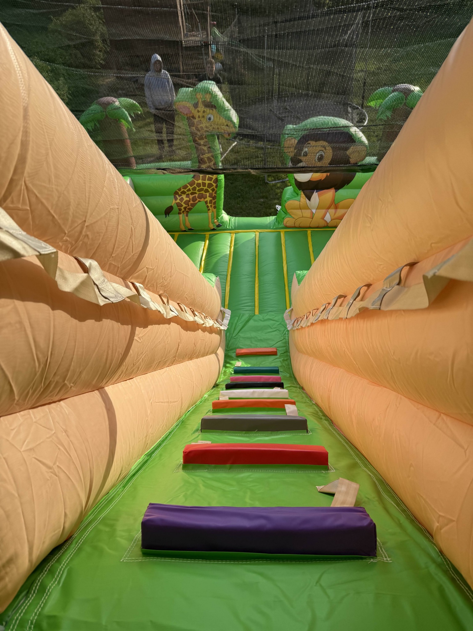 Wnętrze dmuchanej zjeżdżalni z motywami zwierząt, widok z dołu na schody prowadzące na górę. W tle siatka zabezpieczająca i sylwetka osoby.