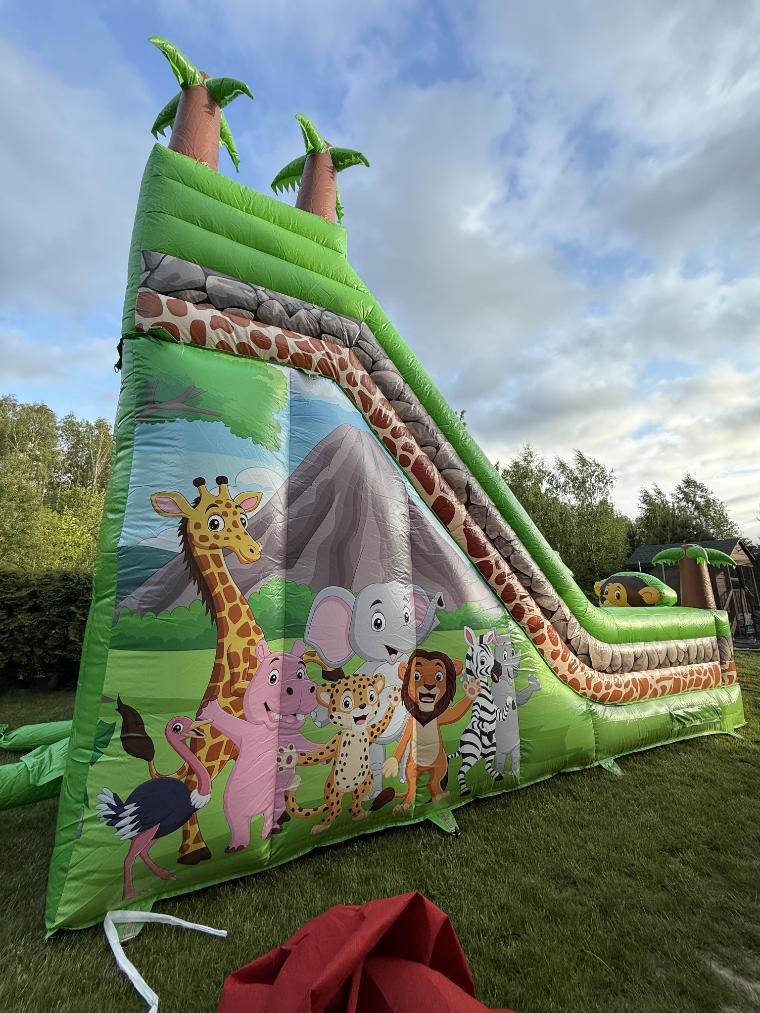 Zielona dmuchana zjeżdżalnia z motywem safari, z palmami na szczycie i wizerunkami zwierząt, ustawiona na trawie w plenerze.