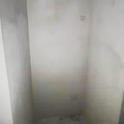 Wnętrze po szpachlowaniu: gładkie, szare ściany z widocznymi puszkami elektrycznymi i fragmentem podłogi. Przygotowanie do malowania, widok na narożnik.