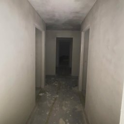 ZIAJBUD - Korytarz po szpachlowaniu: surowe ściany i sufit w odcieniach szarości, widoczne ślady prac wykończeniowych na podłodze, otwarte przejścia drzwiowe.