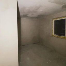 ZIAJBUD - Surowe wnętrze pokoju po szpachlowaniu, z widocznymi śladami prac na ścianach i suficie. Okno z żółtą taśmą zabezpieczającą. Minimalistyczny kadr, neutralne oświetlenie.