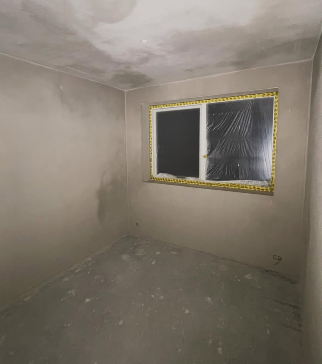 Pokój po szpachlowaniu ścian i sufitu, okno zabezpieczone folią. Widoczne ślady po szpachli na podłodze. Przygotowanie do dalszych prac wykończeniowych.