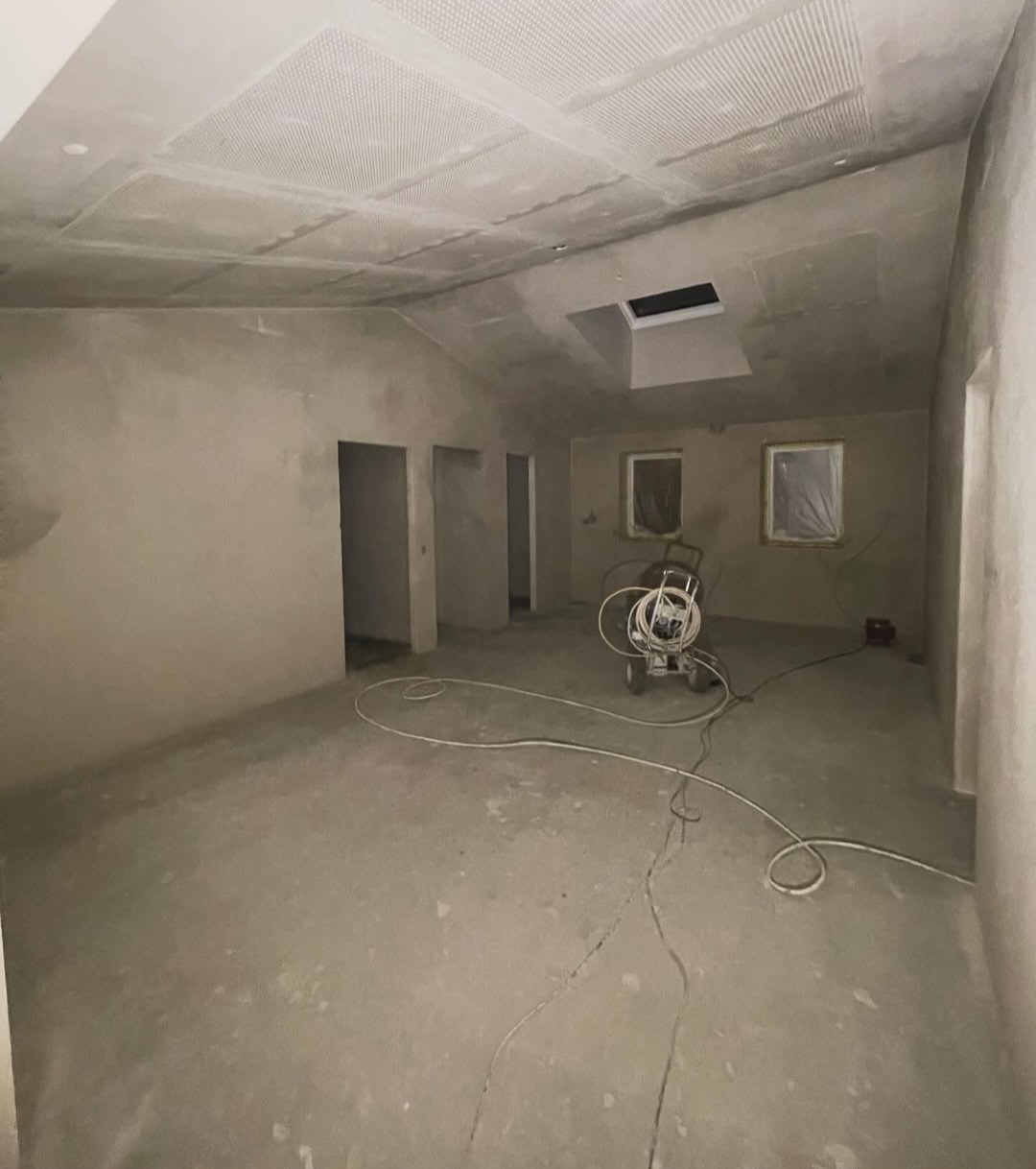 Wnętrze w trakcie szpachlowania: surowe ściany, podłoga i sufit, maszyna do szpachlowania na środku, okna zabezpieczone folią, widoczne kable na podłodze.
