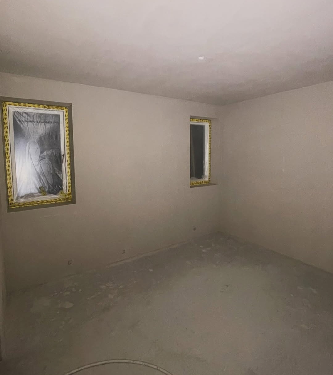 Wnętrze pokoju po szpachlowaniu ścian i sufitu, okna zabezpieczone folią i taśmą malarską, przygotowane do malowania. Minimalistyczny styl.