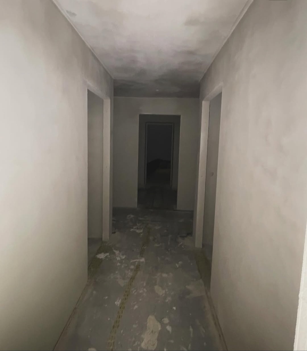 Korytarz po szpachlowaniu: surowe ściany i sufit w odcieniach szarości, widoczne ślady prac wykończeniowych na podłodze, otwarte przejścia drzwiowe.