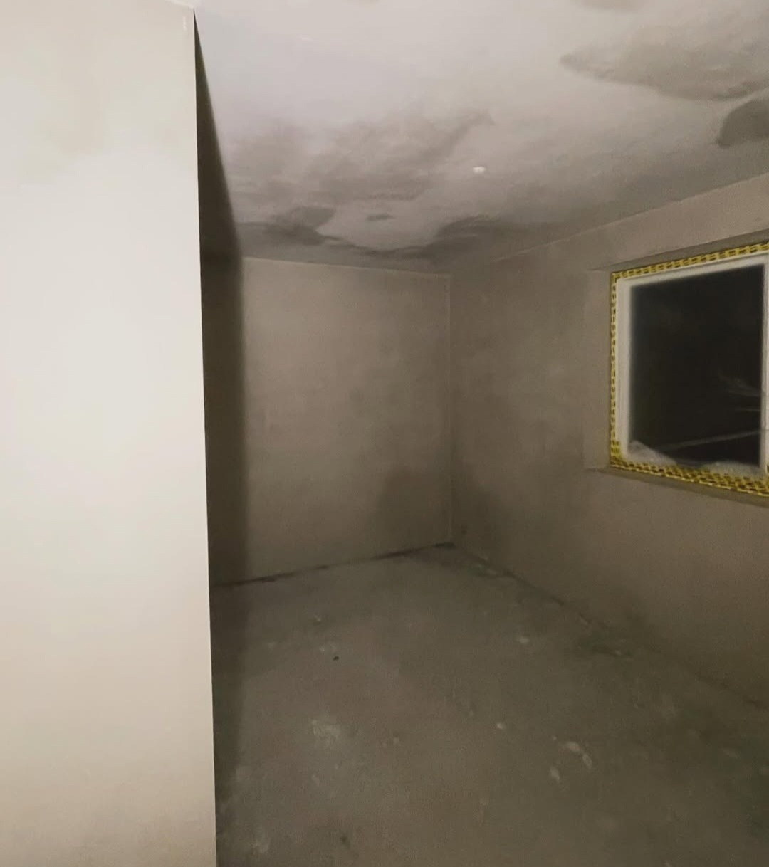 Surowe wnętrze pokoju po szpachlowaniu, z widocznymi śladami prac na ścianach i suficie. Okno z żółtą taśmą zabezpieczającą. Minimalistyczny kadr, neutralne oświetlenie.