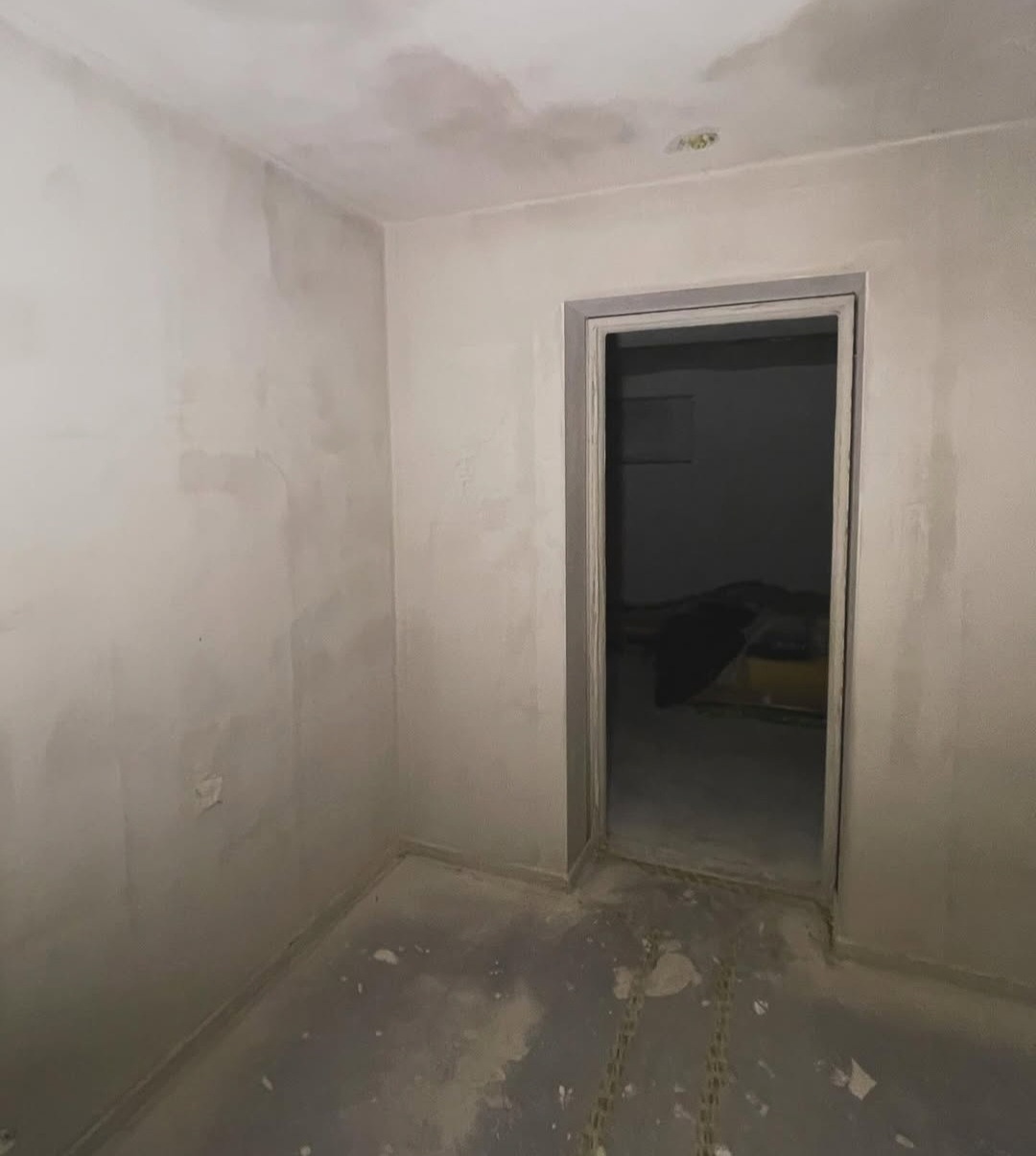 Pokój po szpachlowaniu, przygotowany do malowania. Widoczne ślady szpachli na ścianach i suficie, surowa futryna drzwi. Podłoga wymaga czyszczenia.