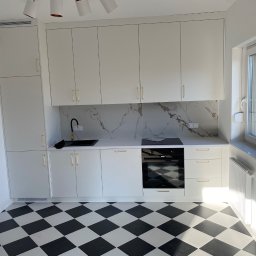 Usługi Ogólnobudowlane Patrycjusz Puzdrowski - Biała kuchnia z czarnym blatem i złotymi uchwytami, wzorzysty panel ścienny imitujący marmur, czarno-biała podłoga w szachownicę, nowoczesny design.