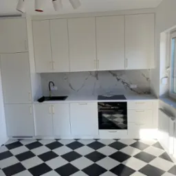 Biała kuchnia z czarnym blatem i złotymi uchwytami, wzorzysty panel ścienny imitujący marmur, czarno-biała podłoga w szachownicę, nowoczesny design.