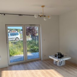 Usługi Ogólnobudowlane Patrycjusz Puzdrowski - Jasny pokój po remoncie z panelami podłogowymi, marmurowym stolikiem i widokiem na zieleń przez duże okno balkonowe. Nowoczesna lampa sufitowa.